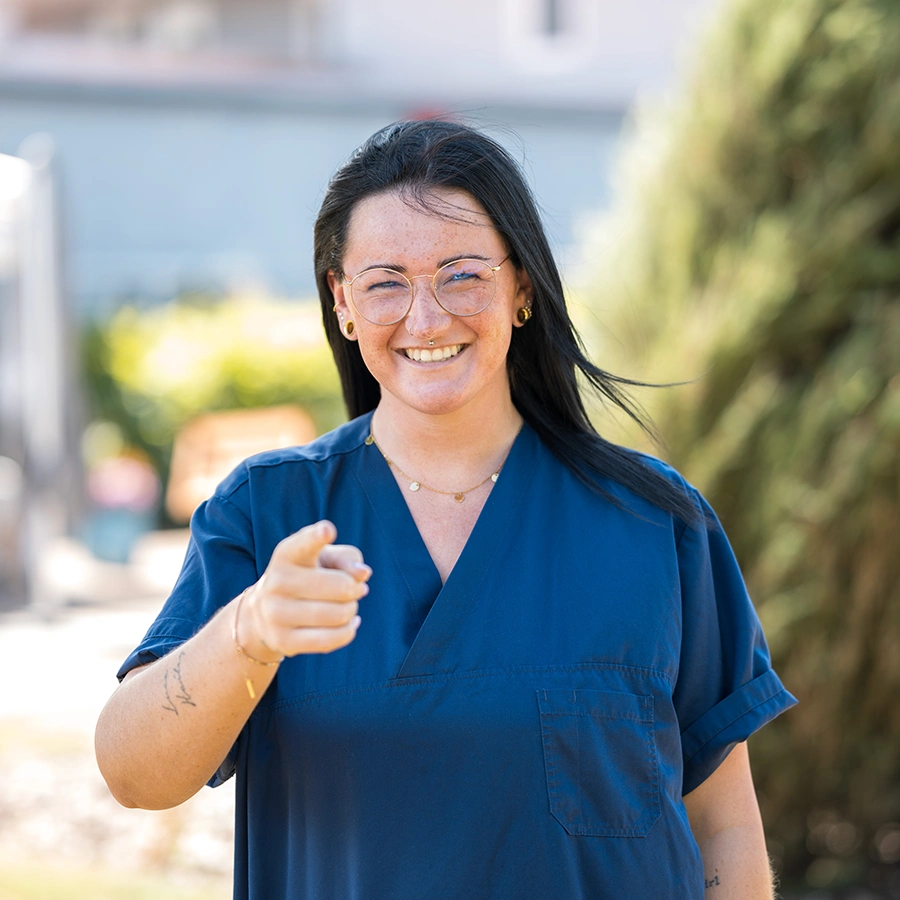 Pflegerin zeigt lächelnd mit dem Zeigefinger einer Hand in die Kamer. Im Hintergrund eine Weg und Busch verschwommen.