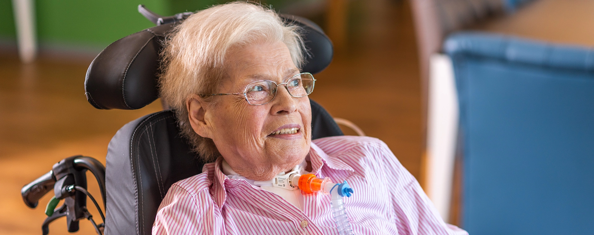 Aufnahme einer Patientin im Rollstuhl schräg von vorne in einem Raum. Sie lächelt in die Ferne.