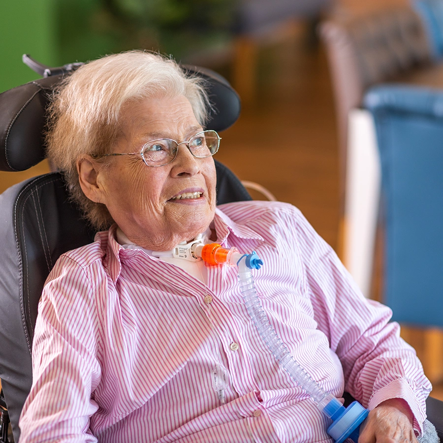 Aufnahme einer Patientin im Rollstuhl schräg von vorne in einem Raum. Sie lächelt in die Ferne.