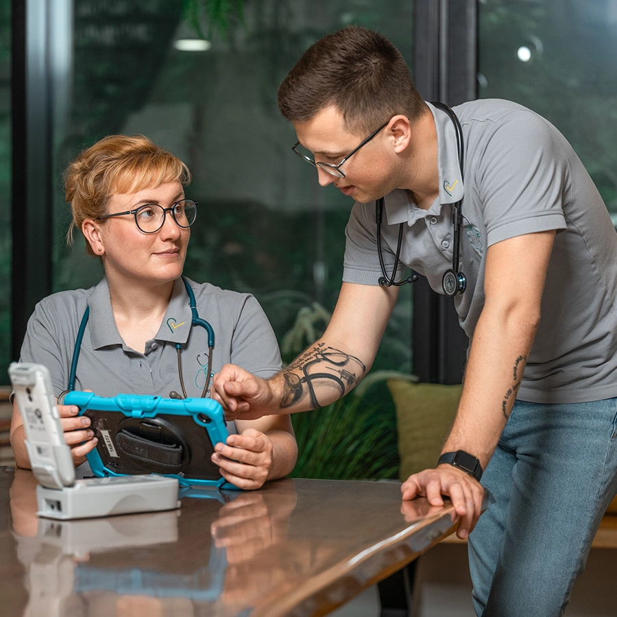Pflegerin sitzt am Tisch mit einem Laptop in der Hand und schaut zu einem Pfleger, der sich vornüber gebeugt auf dem Tisch abstützt und auf das Tablet schaut.
