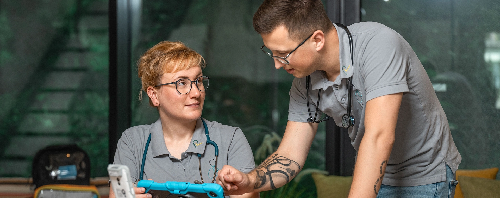 Pflegerin sitzt am Tisch mit einem Laptop in der Hand und schaut zu einem Pfleger, der sich vornüber gebeugt auf dem Tisch abstützt und auf das Tablet schaut.