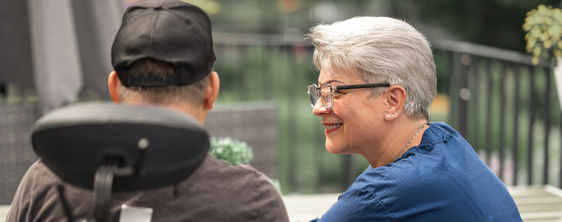Weibliche Pflegekraft in Seitenansicht kniet an einer Seite eines Rollstuhls, auf dem ein Klient sitzt, der von hinten zu sehen ist. Im Hintergrund verschwommen eine Terrasse mit Metallgeländer.